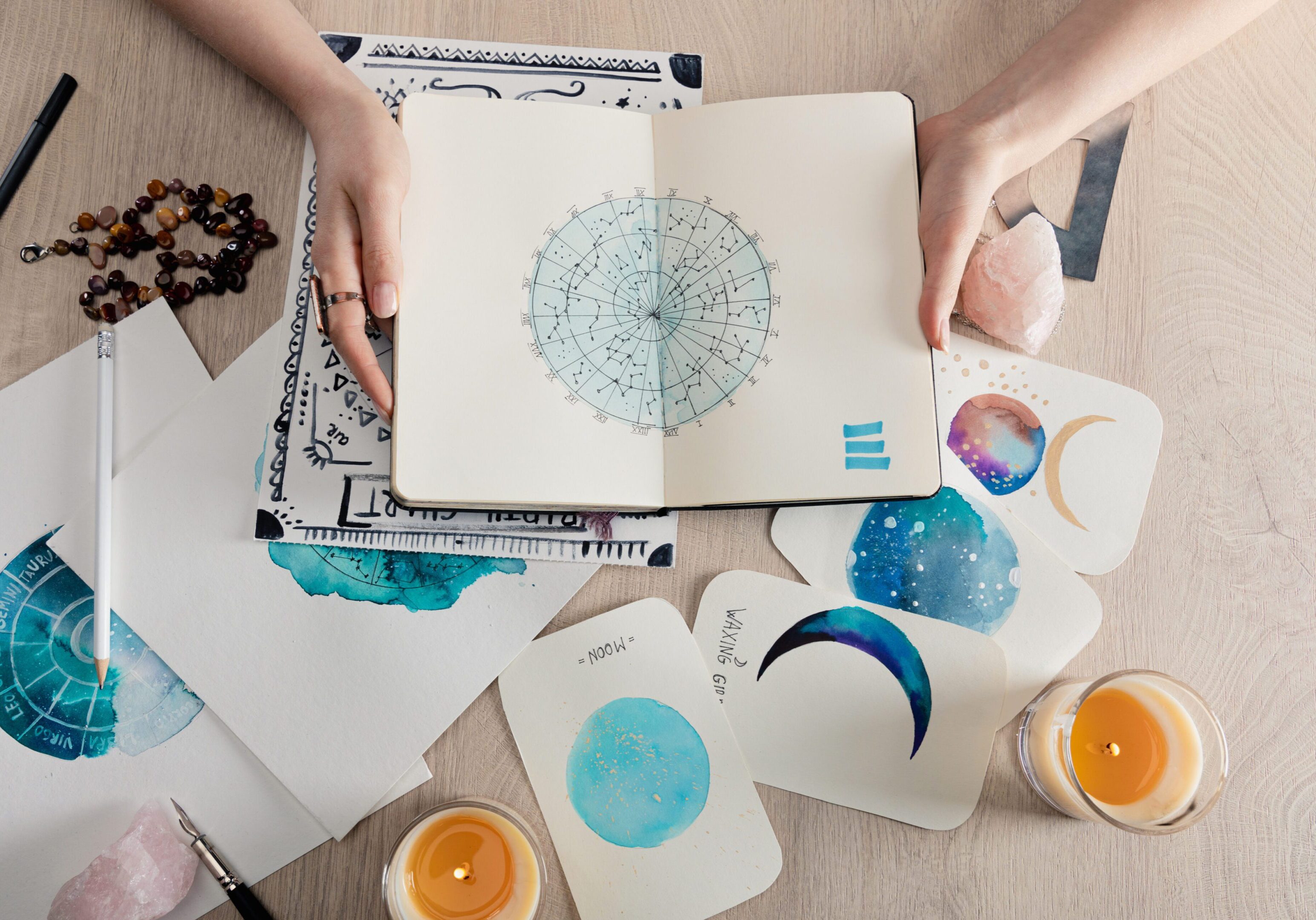 Top view of astrologer holding notebook with watercolor drawings and zodiac signs on cards on table